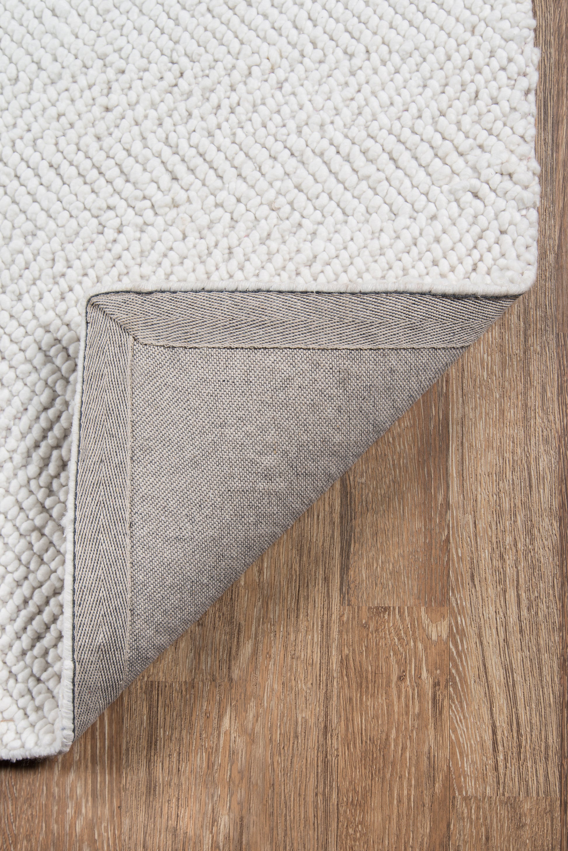 Ivory loop-pile rug with textured surface, folded to reveal gray woven underside and striped binding on wood floor.