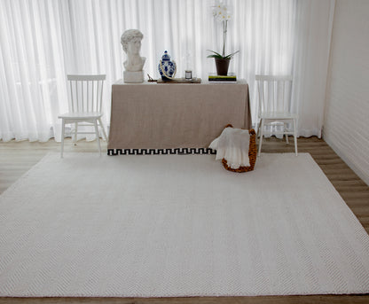 Beige rug with a black Greek-key border on the floor; white chairs, a bust statue, blue vase, orchid, and books nearby.