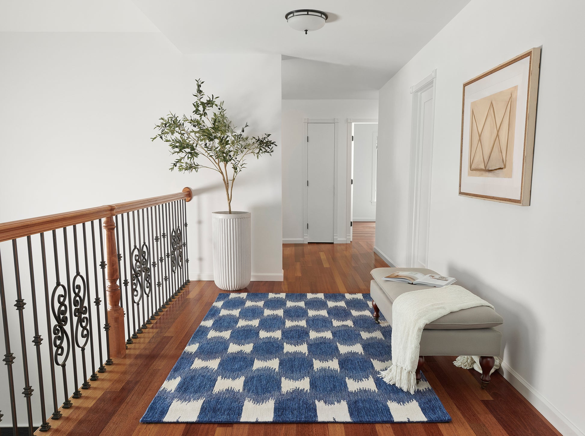 Bright foyer landing with wooden railing, iron balusters, blue-white geometric rug, gray bench with cream throw, potted tree.
