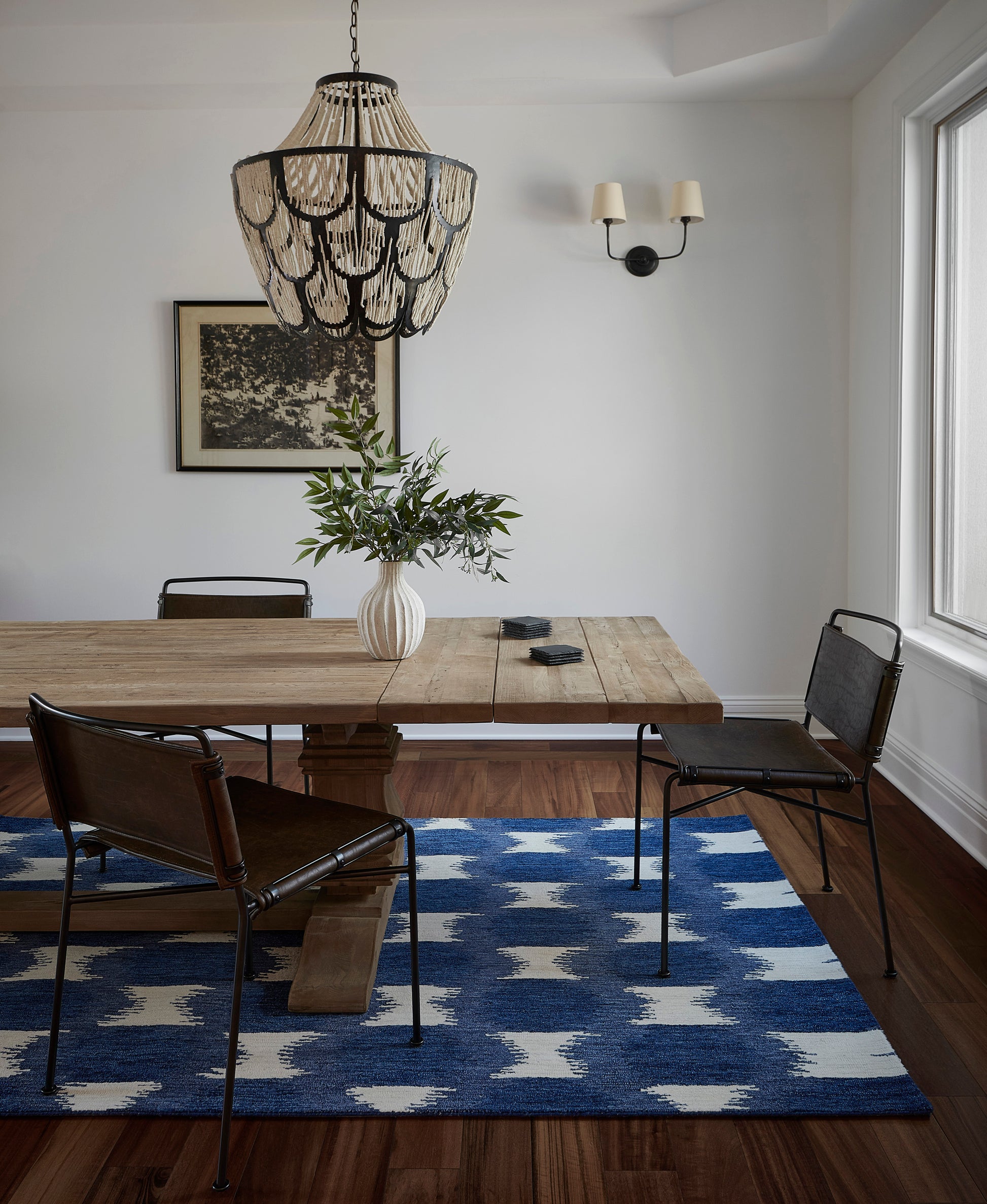 Rustic wooden dining table with black metal chairs, white vase of greenery, blue geometric rug, woven chandelier.