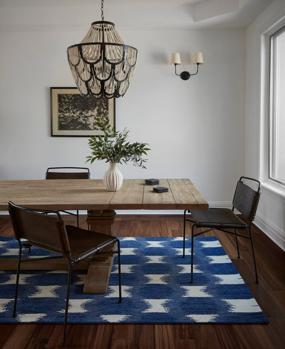 Rustic wooden dining table with black metal chairs, white vase of greenery, blue geometric rug, woven chandelier.