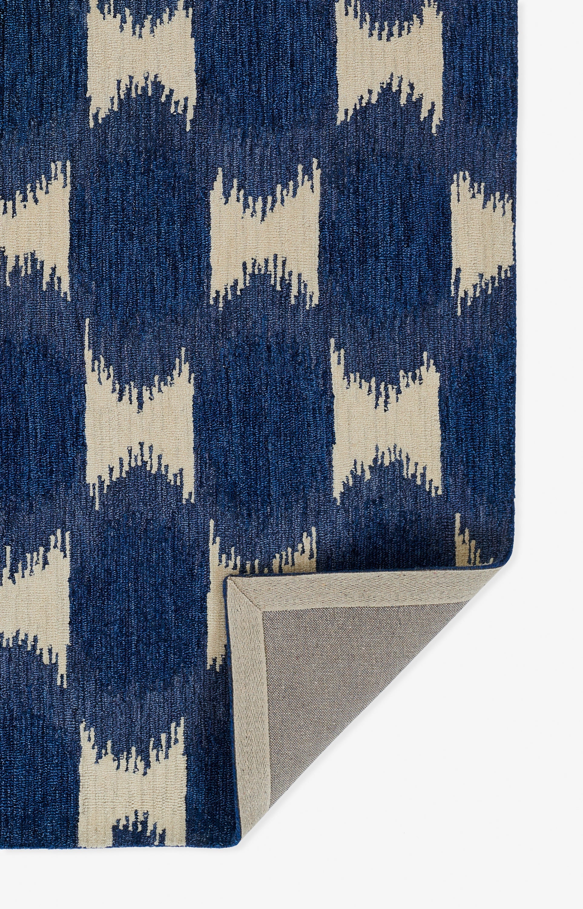Blue and cream ikat-style woven rug with repeating geometric blocks; folded corner shows gray underside.