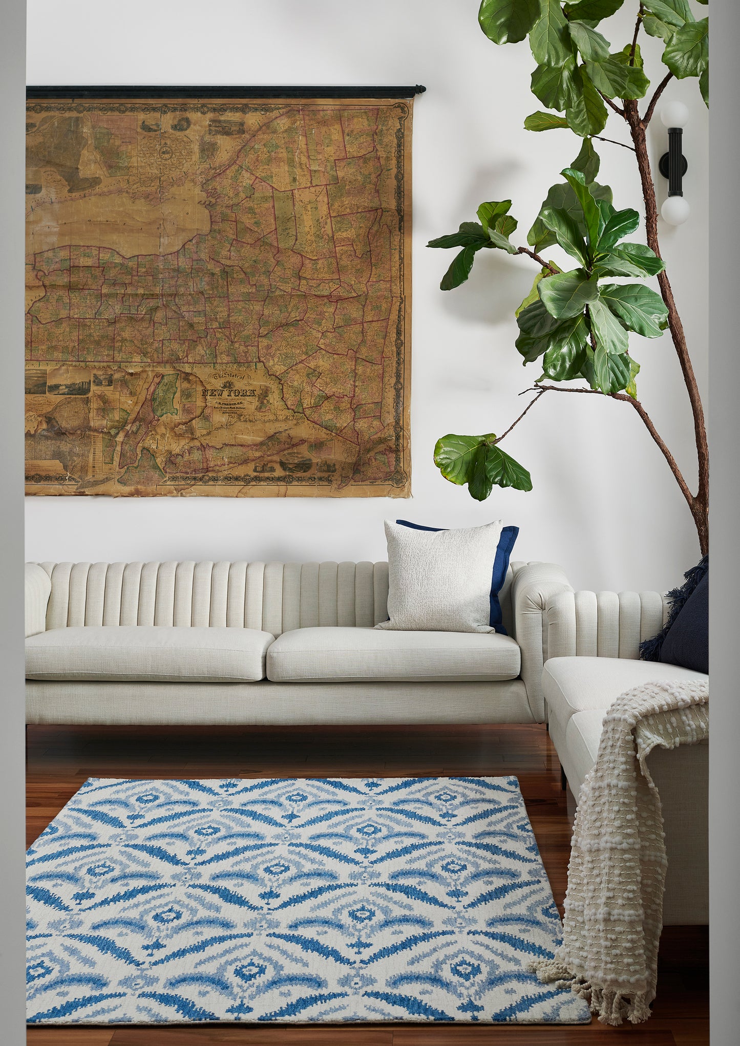 Cozy living room with vintage map wall hanging, cream ribbed sofa, blue-and-white patterned rug, and a tall green plant.