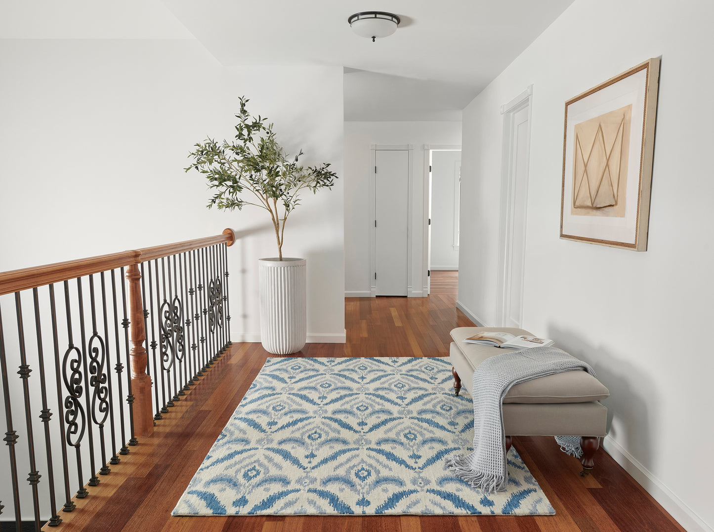 Hallway with blue & white patterned rug, hardwood floor, beige bench with gray throw, potted tree, wall art, iron railing.