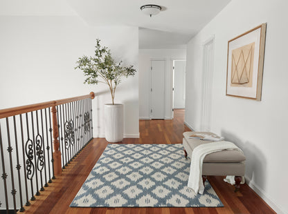 Bright hallway with blue geometric rug, beige upholstered bench with knit throw, potted tree in tall vase, iron railing.