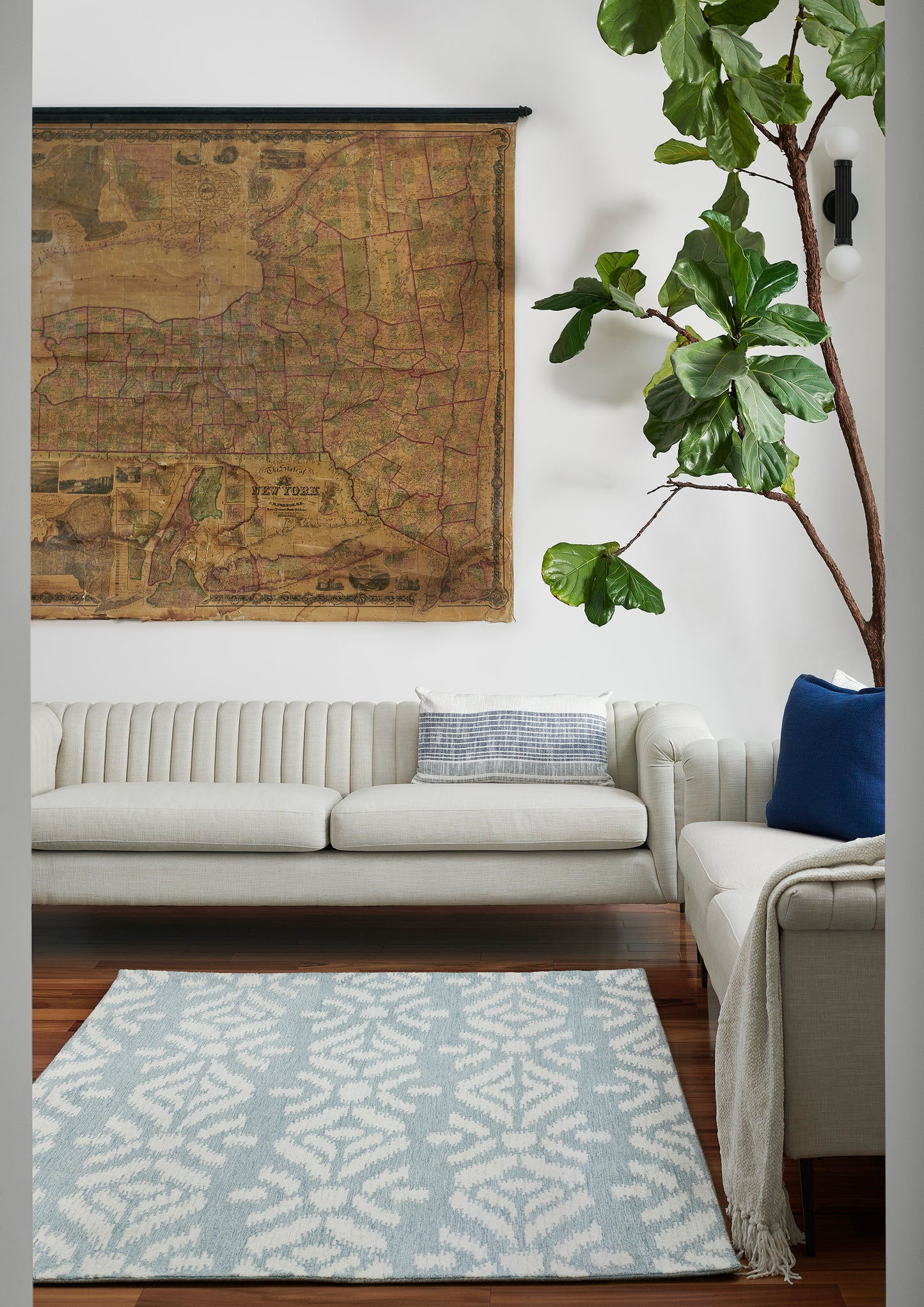 Cozy living room with cream sofa, vintage map wall art, tall fiddle-leaf fig, pale blue patterned rug, blue cushions.
