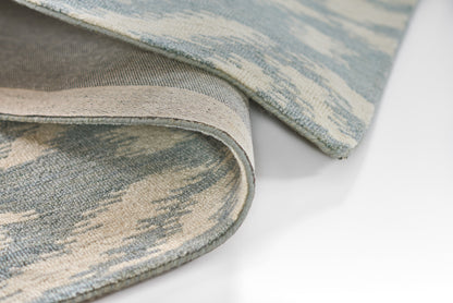 Blue and ivory woven rug with abstract pattern; rolled edge reveals beige backing and textured surface.