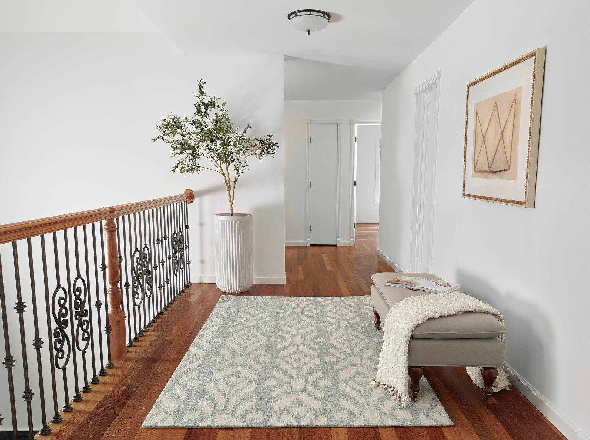 Bright hallway landing with gray patterned rug, wood floor, iron railing, gray bench with throw, tall plant, framed art.