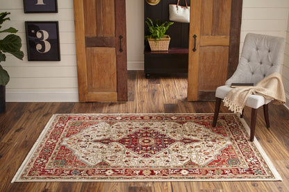 Rectangular oriental rug, 2'X3', in red, ivory and beige on dark wood floor, beside a gray upholstered chair and open wooden doors.