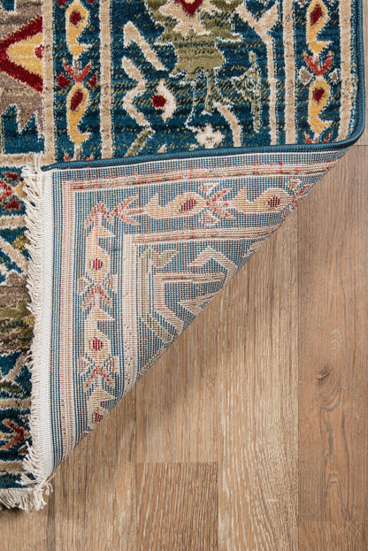 Blue and ivory oriental rug with red and green accents, fringed edge, folded back to show underside on a wooden floor.