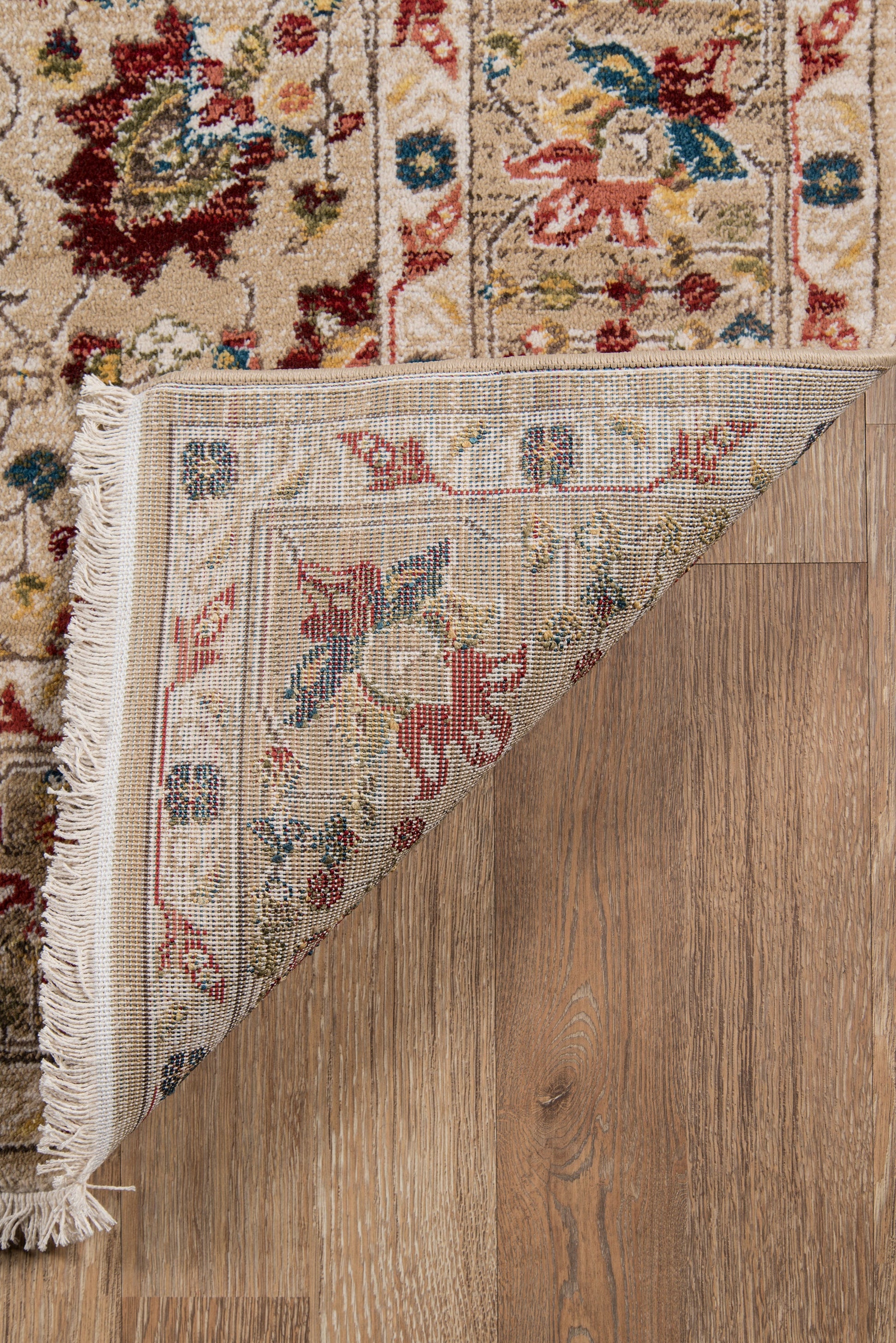 Ivory oriental rug with blue and red floral motifs and fringe; folded corner reveals underside on a wood floor.