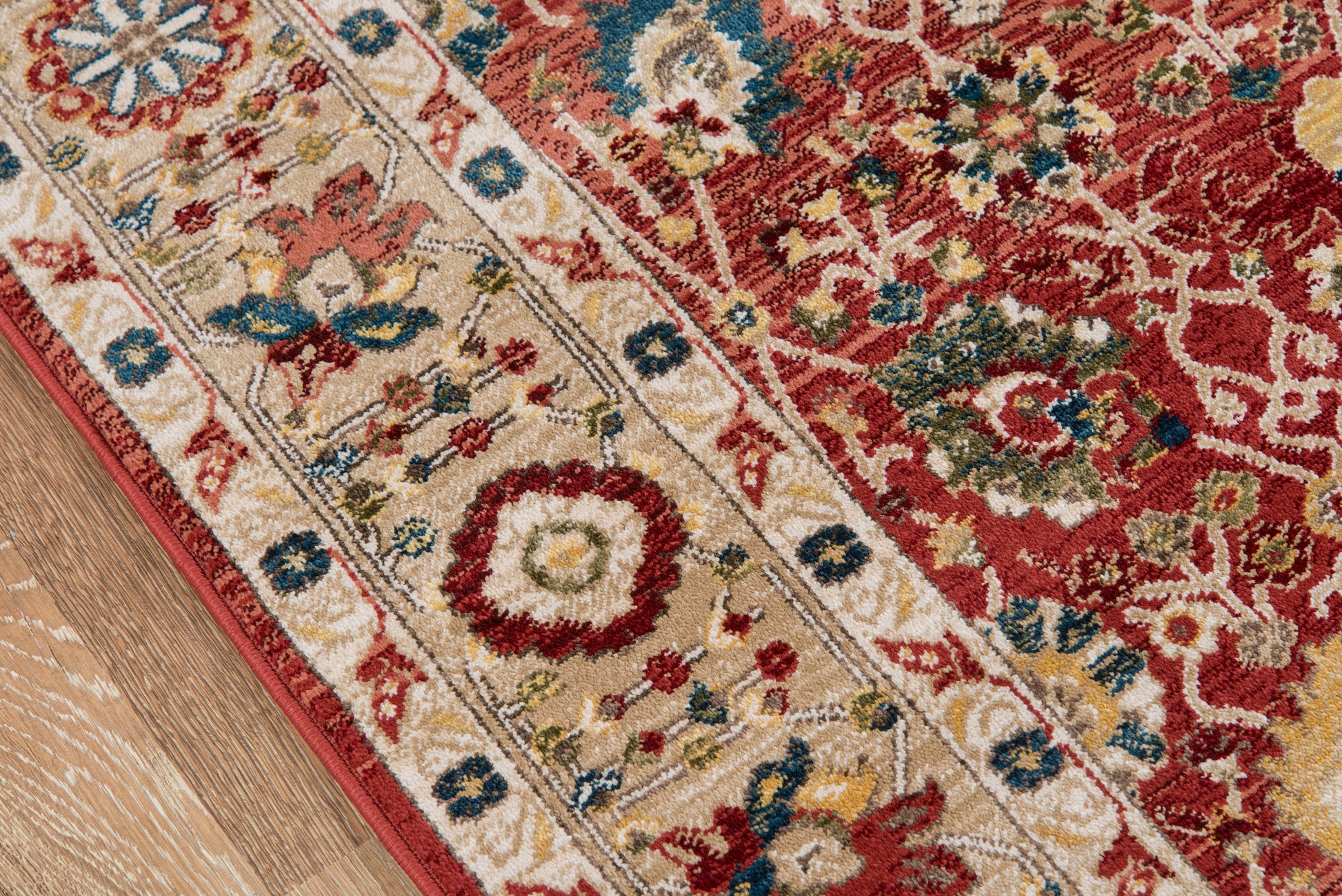 Traditional oriental rug on wooden floor; red field with floral motifs and a beige border with blue accents.