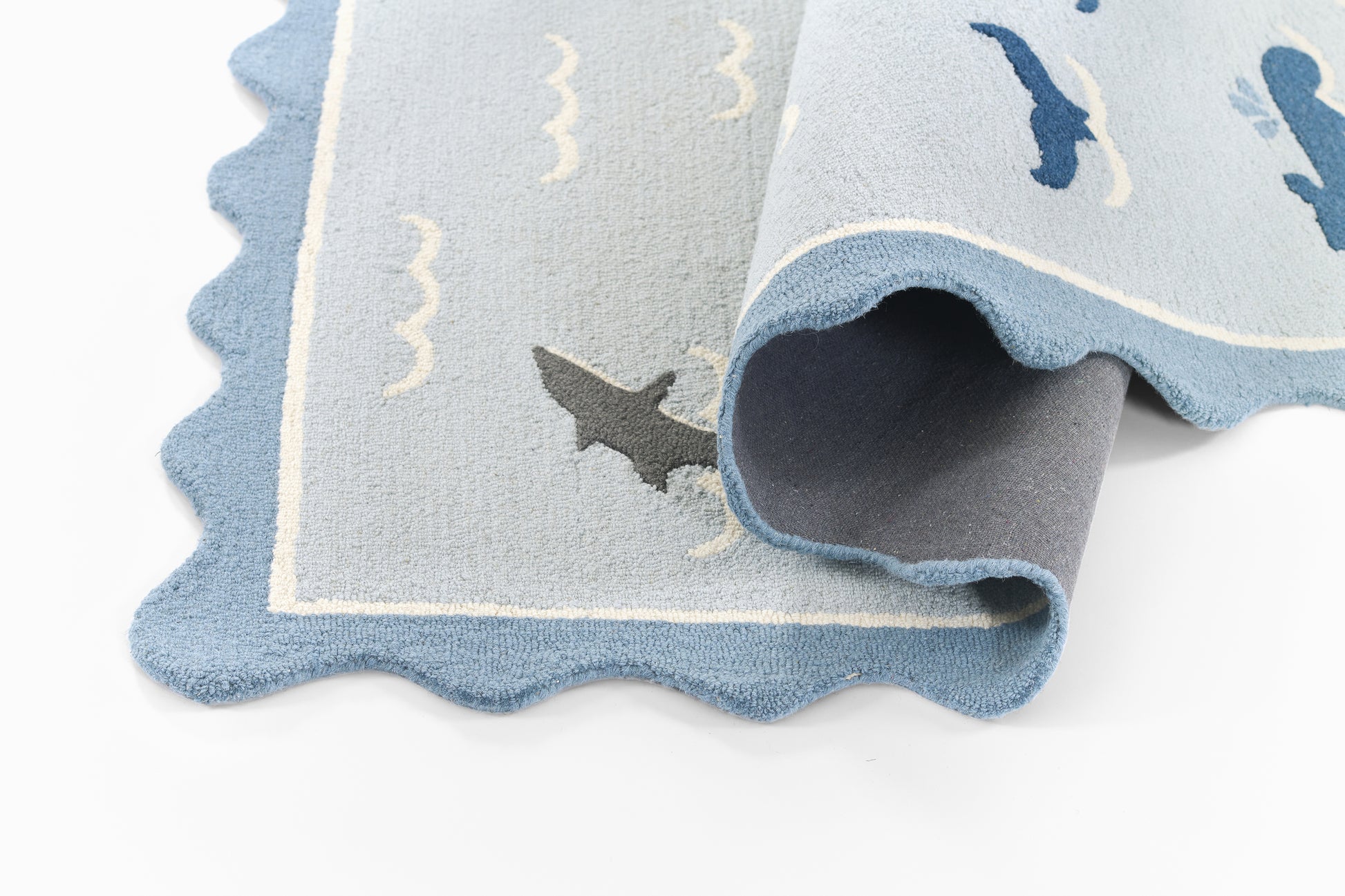 Light blue plush rug with scalloped edges and darker blue trim, featuring shark silhouettes and wave motifs for kids' rooms.