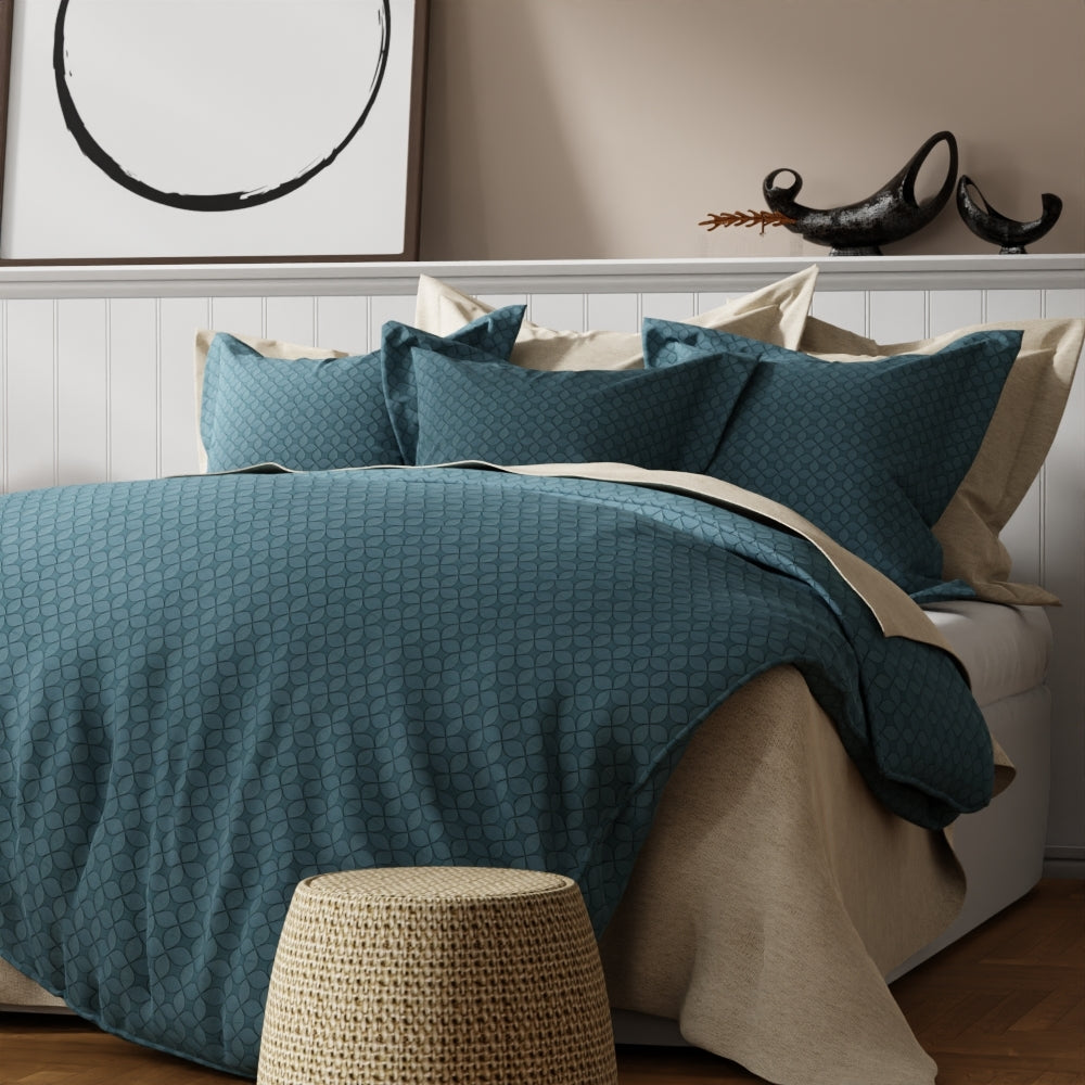 Teal geometric duvet set with matching shams on a beige bed, beige throw, and a woven wicker stool against a paneled wall.