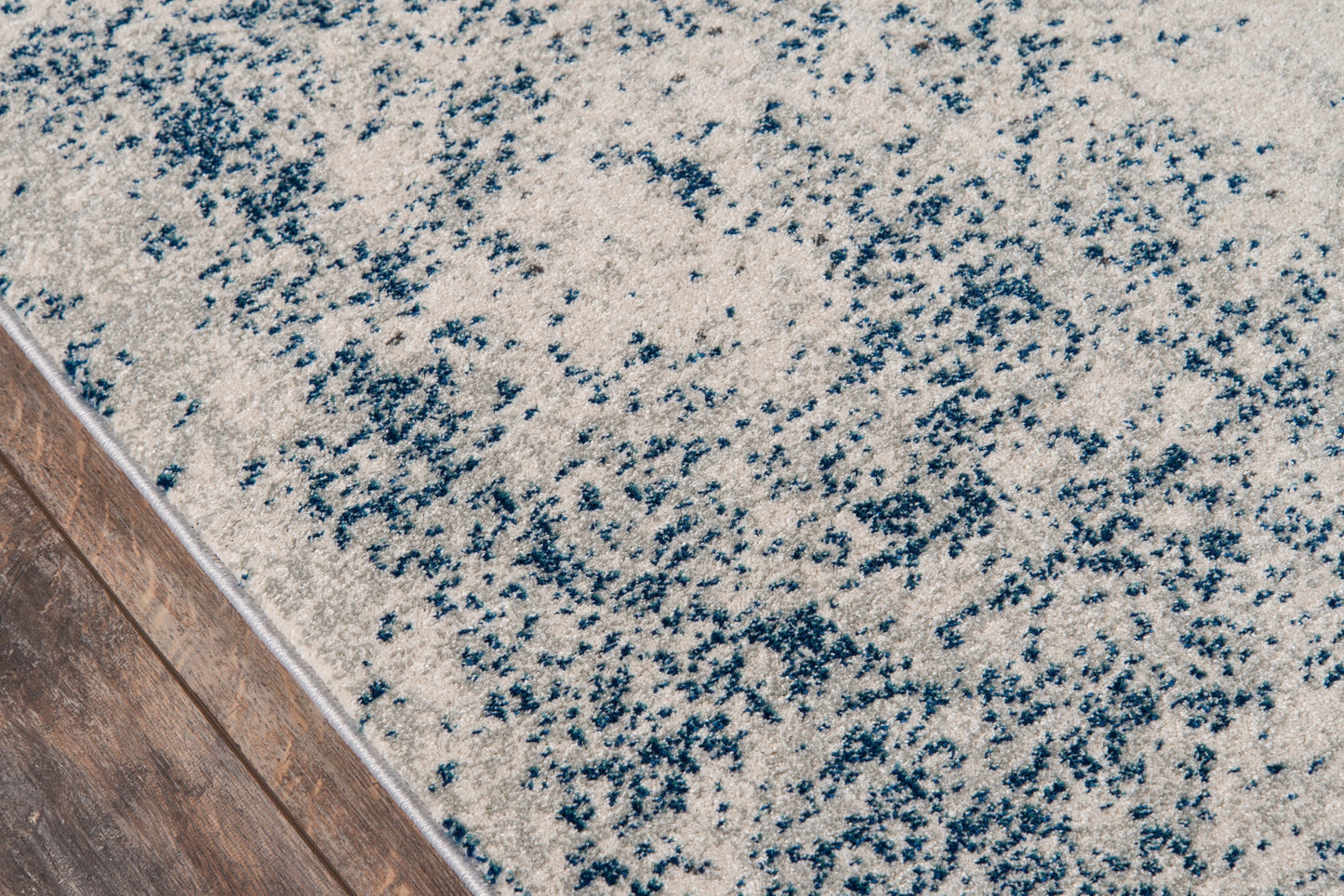 Beige rug with blue speckled abstract pattern and plush pile beside wooden floor edge.