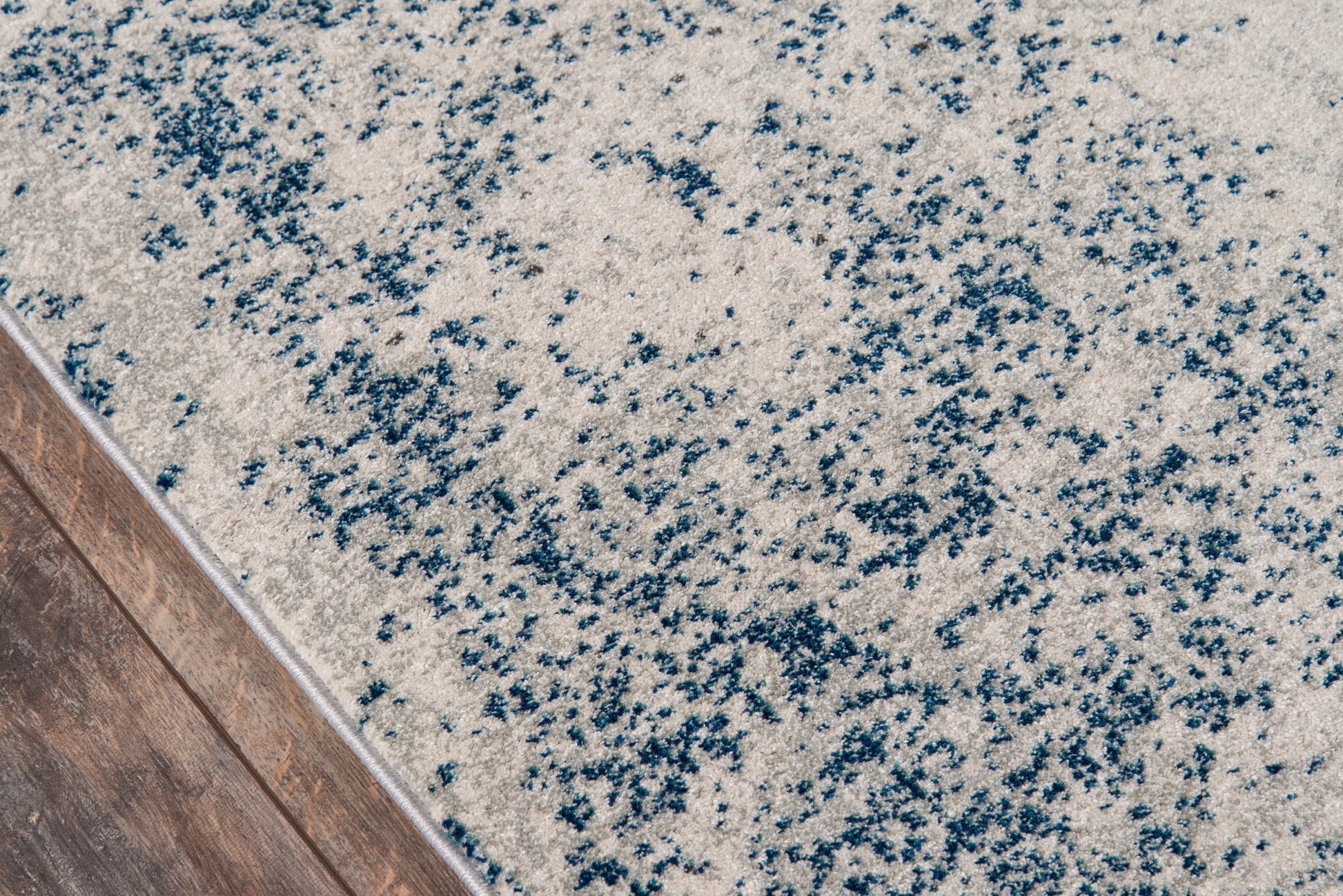 Beige rug with blue speckled abstract pattern and plush pile beside wooden floor edge.