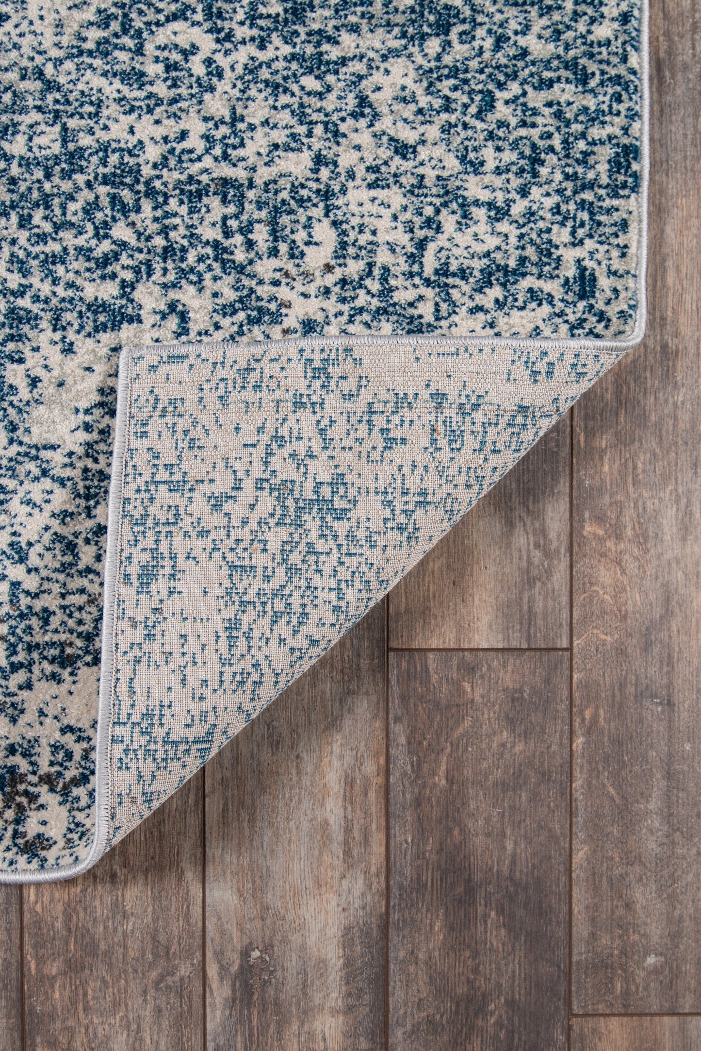 Rectangular blue and white speckled area rug with light underside folded at corner on wood floor.