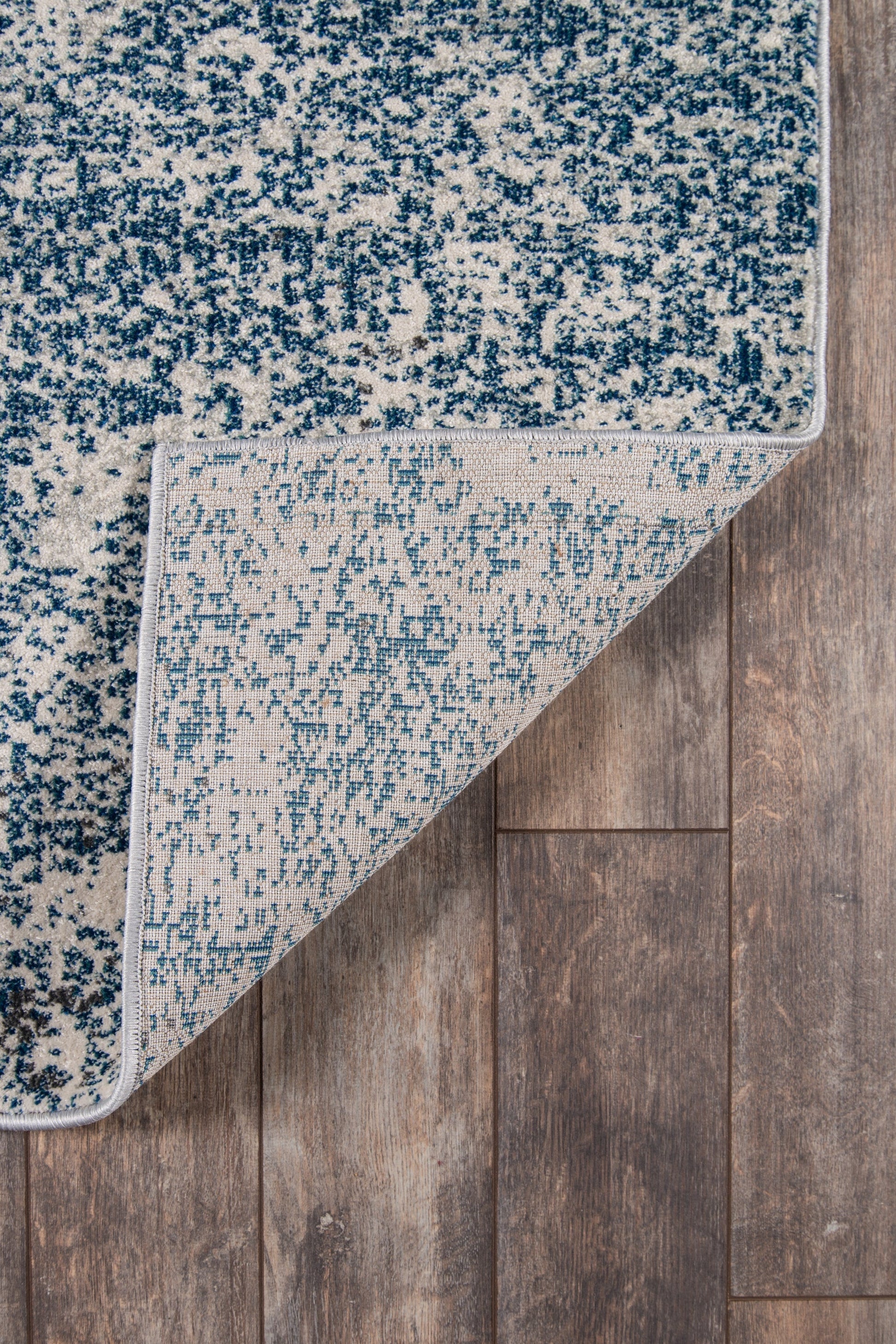 Rectangular blue and white speckled area rug with light underside folded at corner on wood floor.