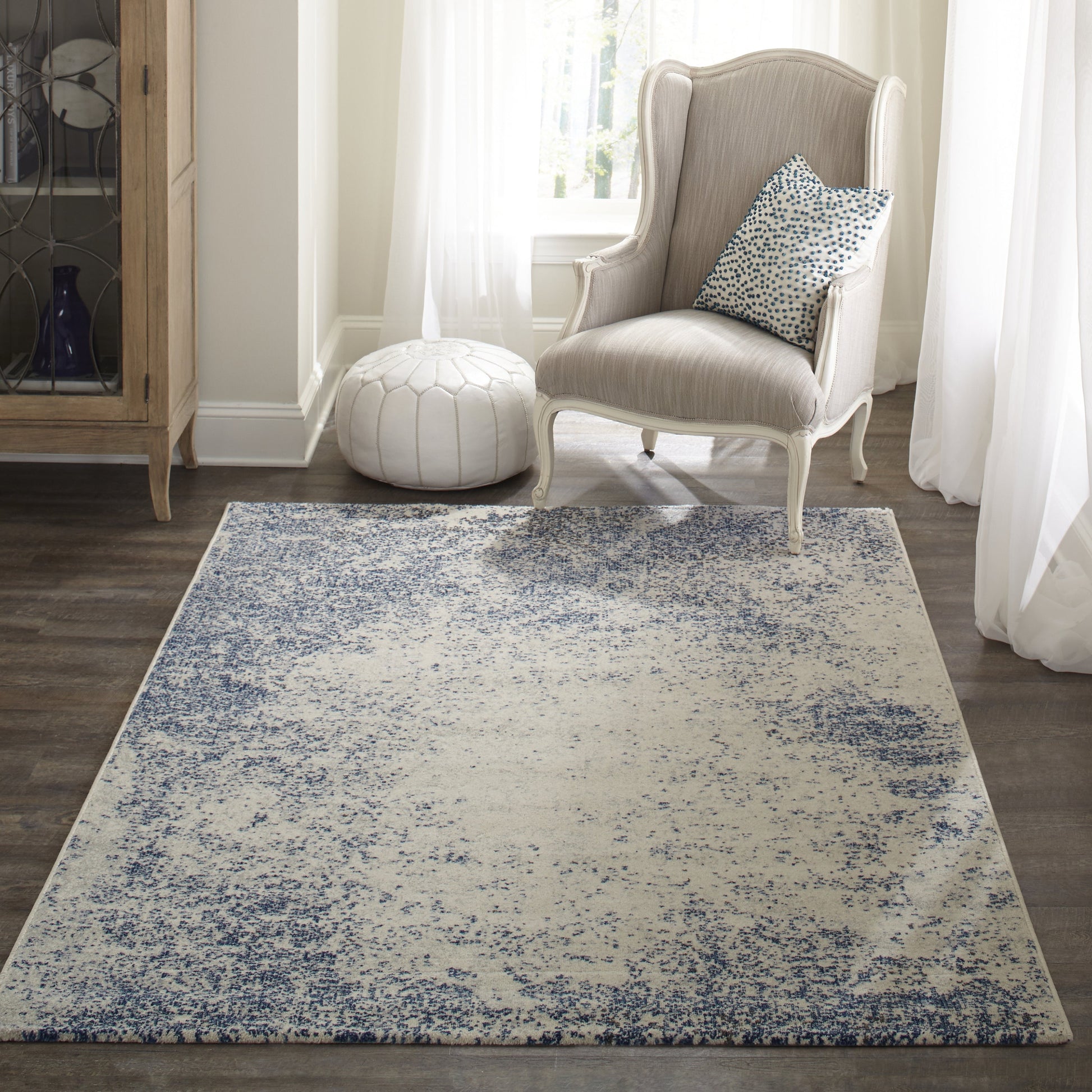 Blue and ivory distressed-pattern rectangular rug, low-pile, in a light-filled living room with a beige wingback chair and white pouf.