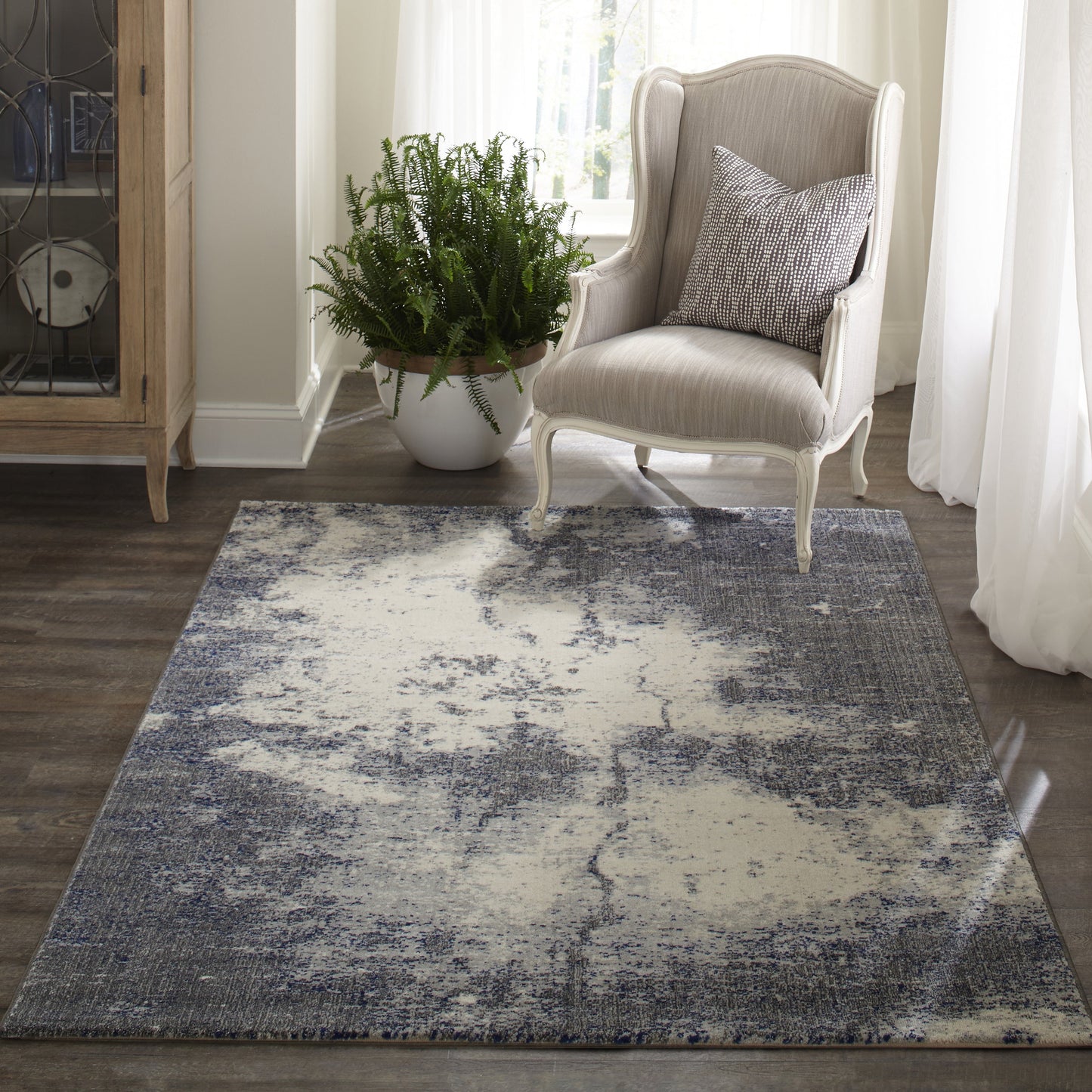 Abstract gray and cream distressed area rug on wood floor beside a light upholstered armchair and plant.