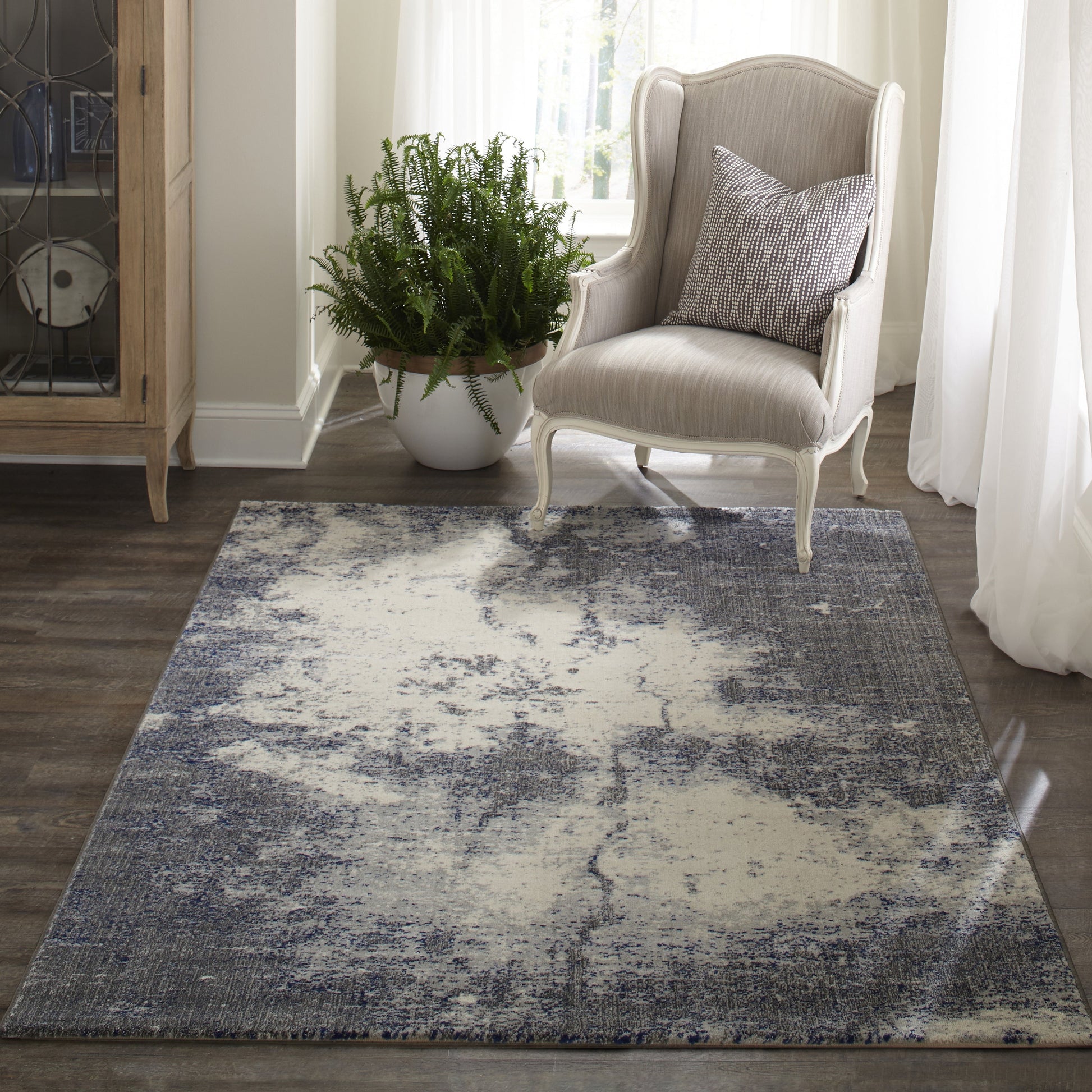 Abstract gray and cream distressed area rug on wood floor beside a light upholstered armchair and plant.