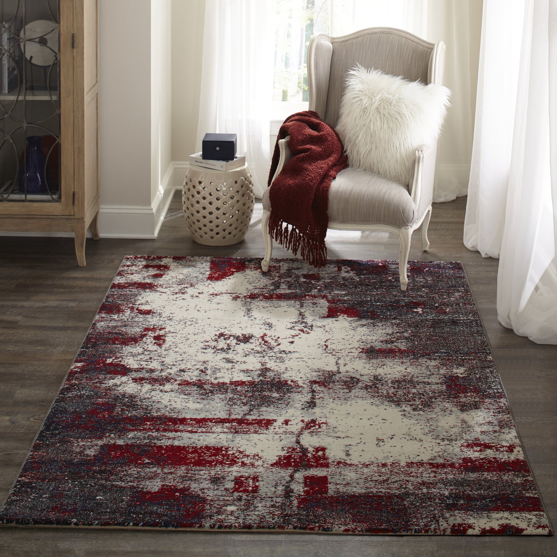 Abstract distressed rug in red, gray, and ivory beside a gray upholstered armchair with a red throw and white fluffy pillow.
