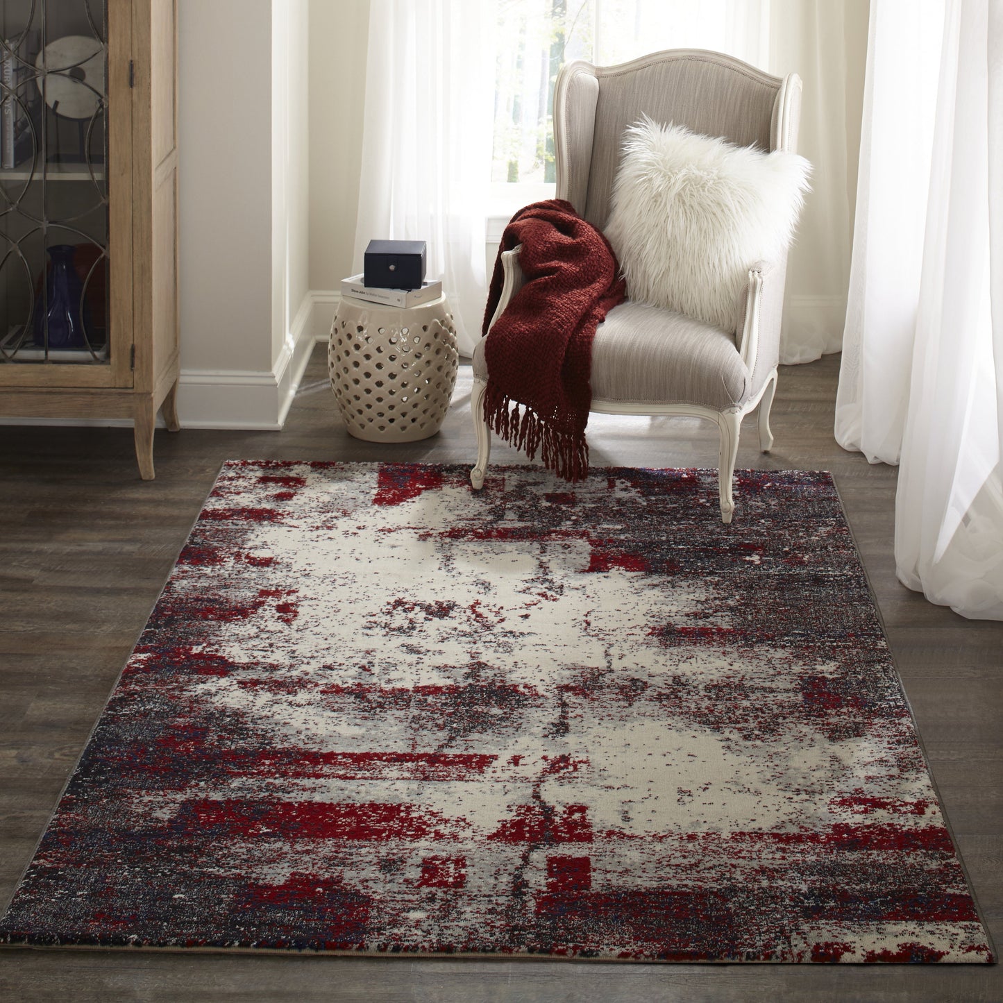 Abstract rug in gray, ivory and burgundy under a tufted light-gray armchair with white fur pillow and burgundy throw.