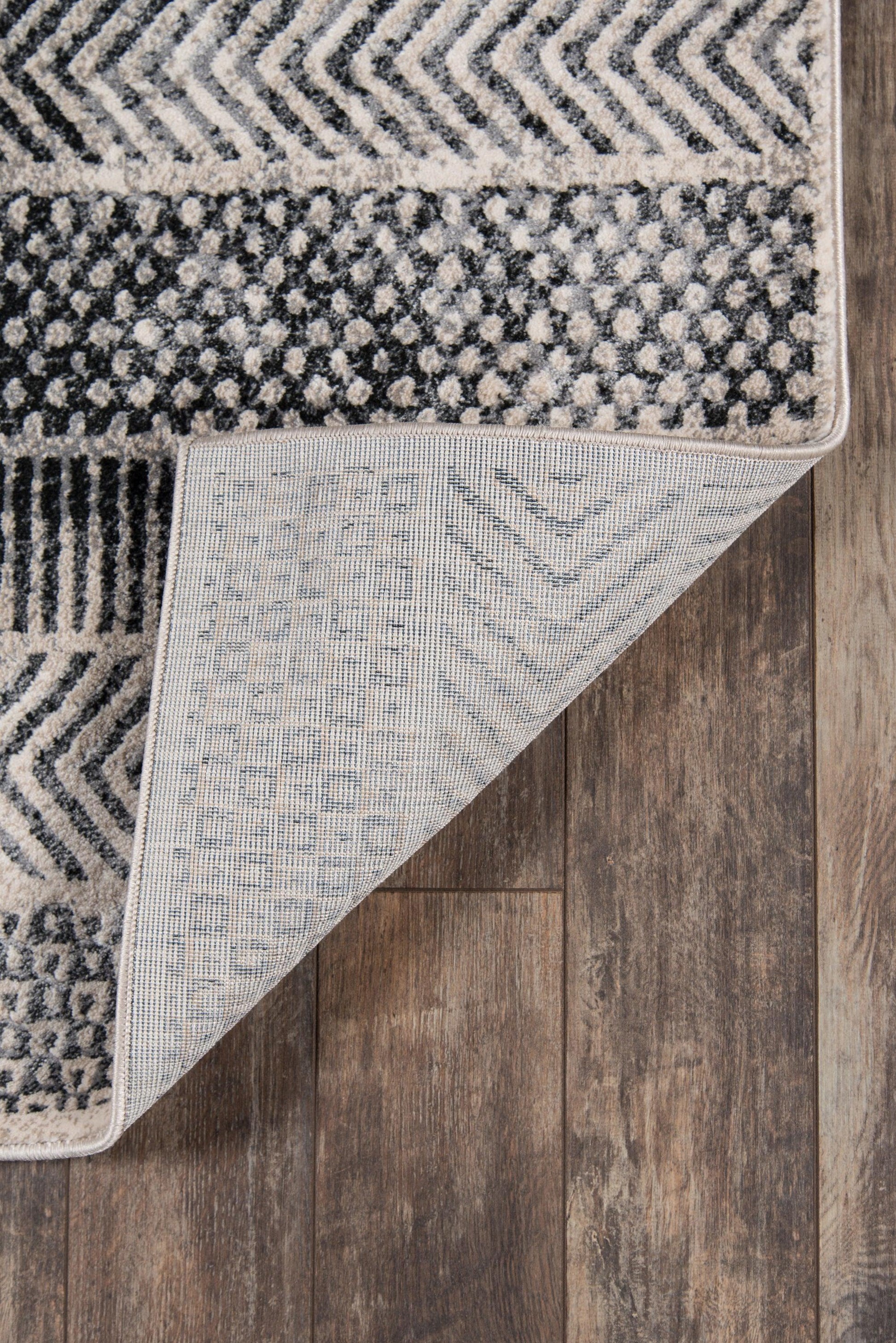 Gray geometric area rug with chevron and dot patterns, folded corner shows lighter underside, charcoal ivory hues.