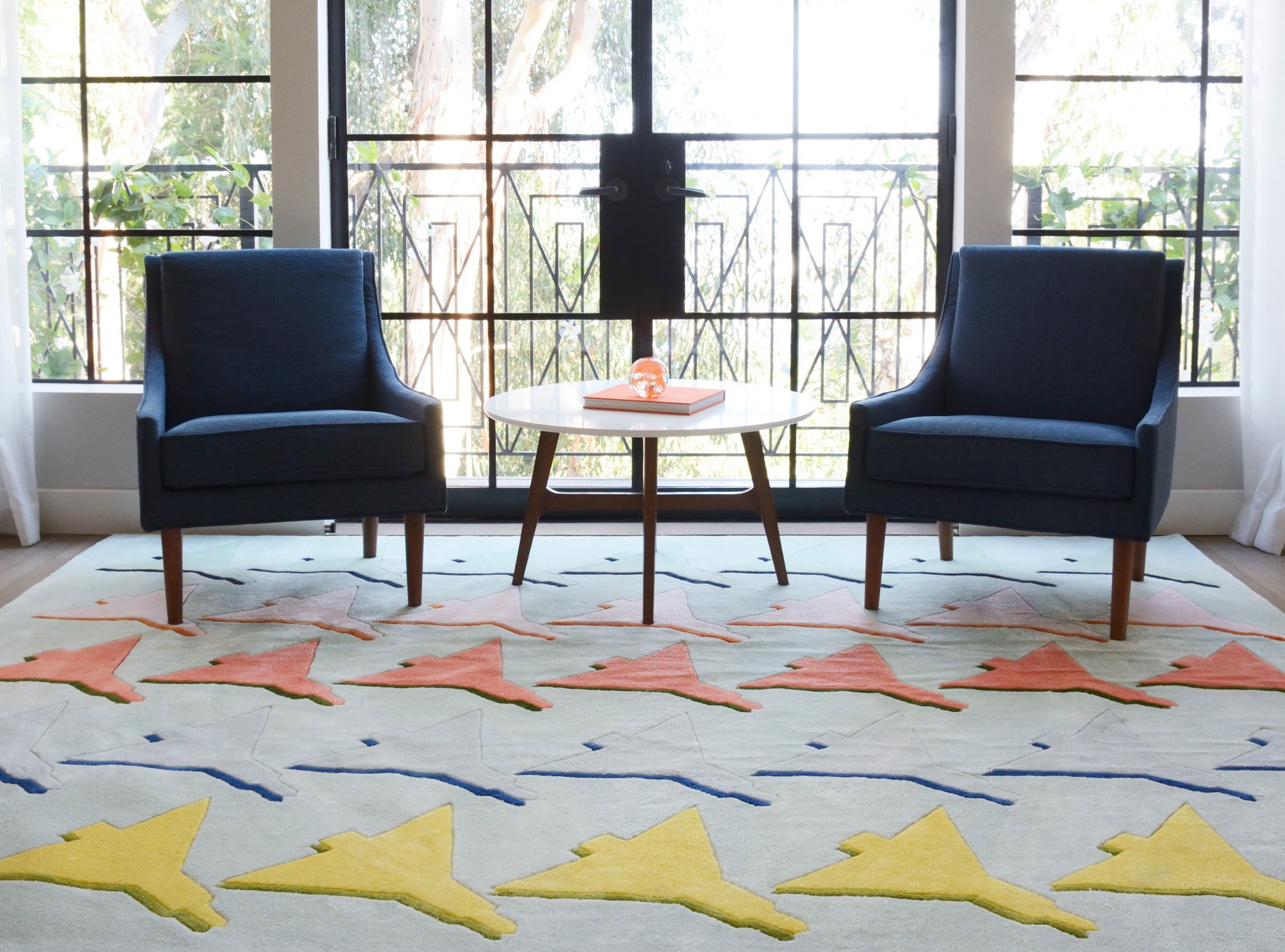 Colorful fish-pattern rug on the floor in a sunlit living room, flanked by two navy blue mid-century armchairs & a round coffee table.