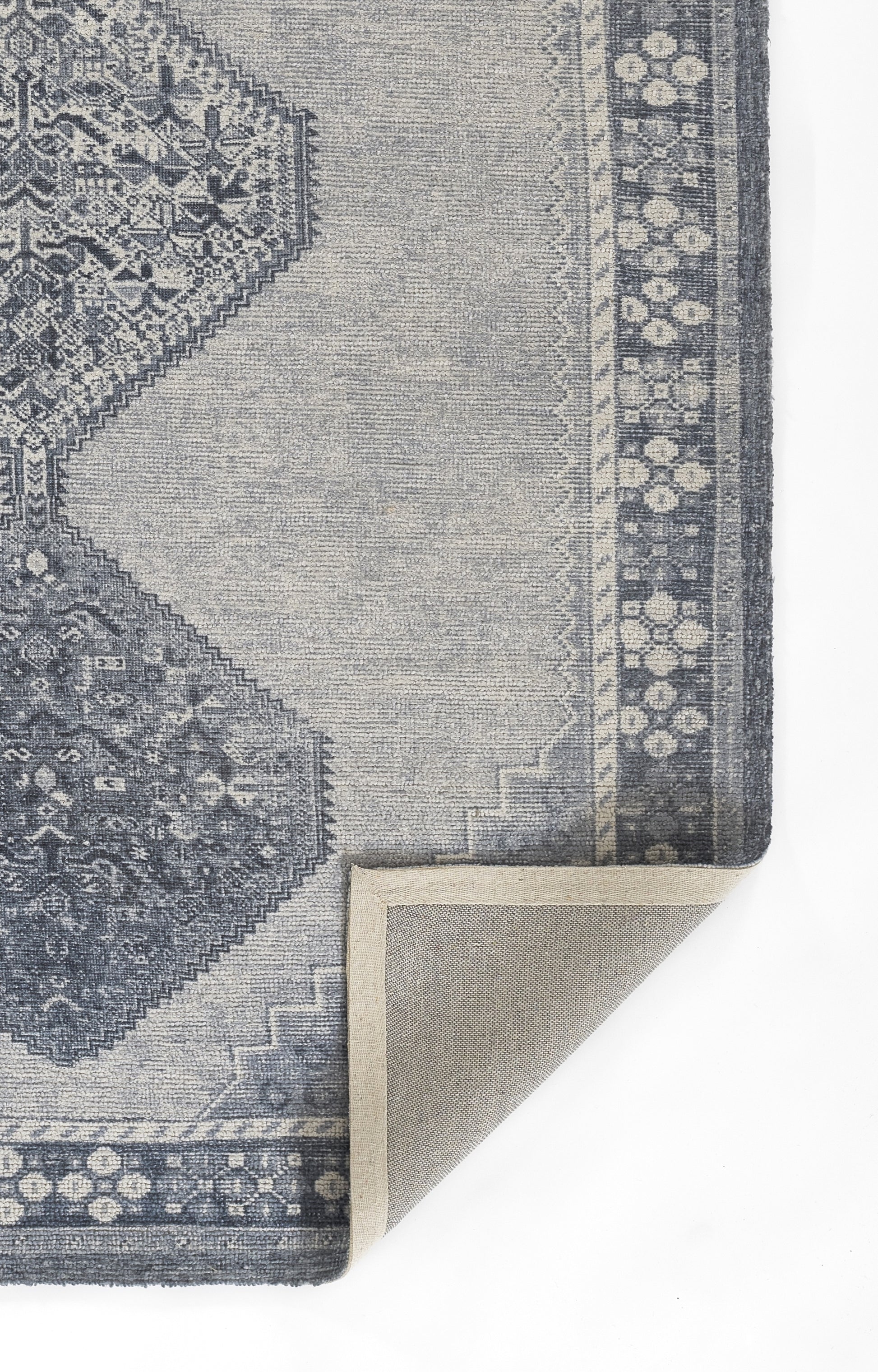Blue-gray woven rug with ornate medallion and border; corner folded to reveal tan backing.
