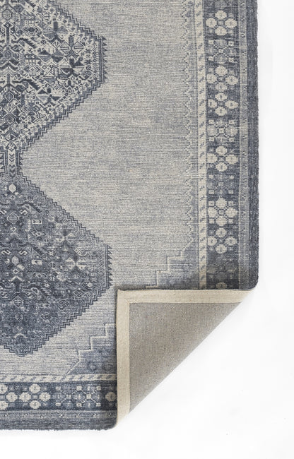 Blue-gray patterned rug with geometric medallions and decorative border; fold reveals beige underside.