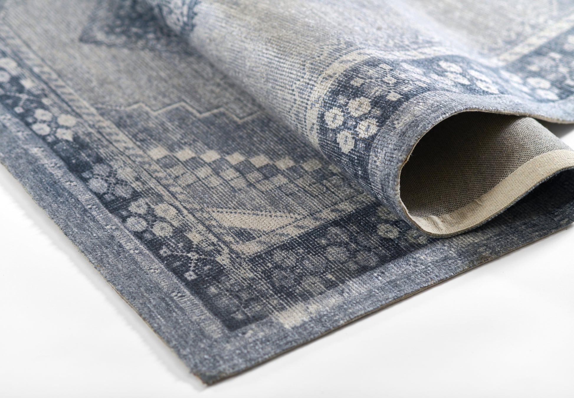 Blue-gray patterned rug with vintage geometric motifs, woven texture; folded corner reveals beige underside.