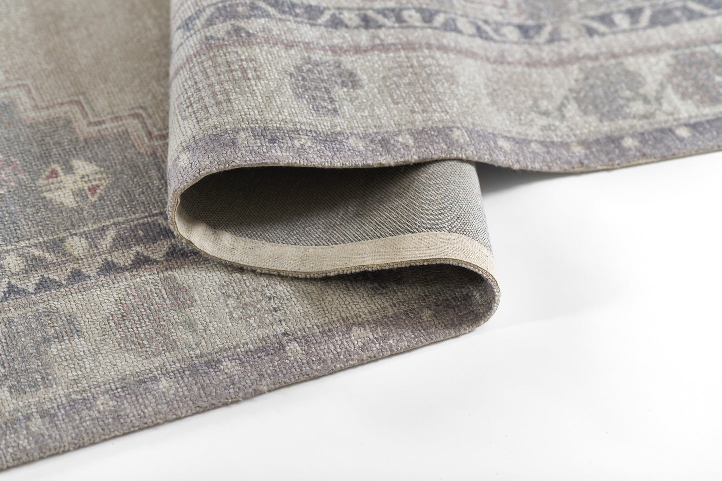 Muted gray-lavender woven rug with subtle geometric motifs; rolled edge reveals a beige underside.
