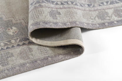 Muted gray-lavender woven rug with subtle geometric motifs; rolled edge reveals a beige underside.
