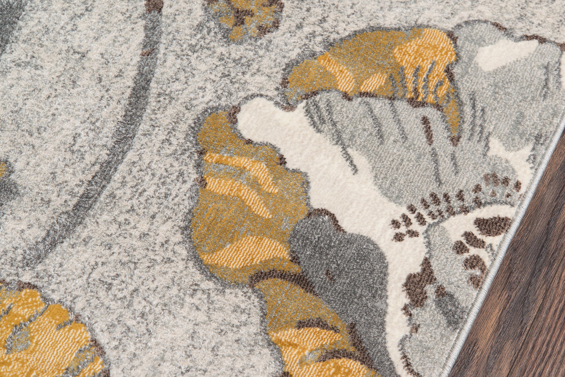 Close-up of gray and gold floral-patterned rug on light beige backdrop, edge resting on dark wood floor.