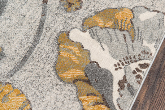 Close-up of gray and gold floral-patterned rug on light beige backdrop, edge resting on dark wood floor.