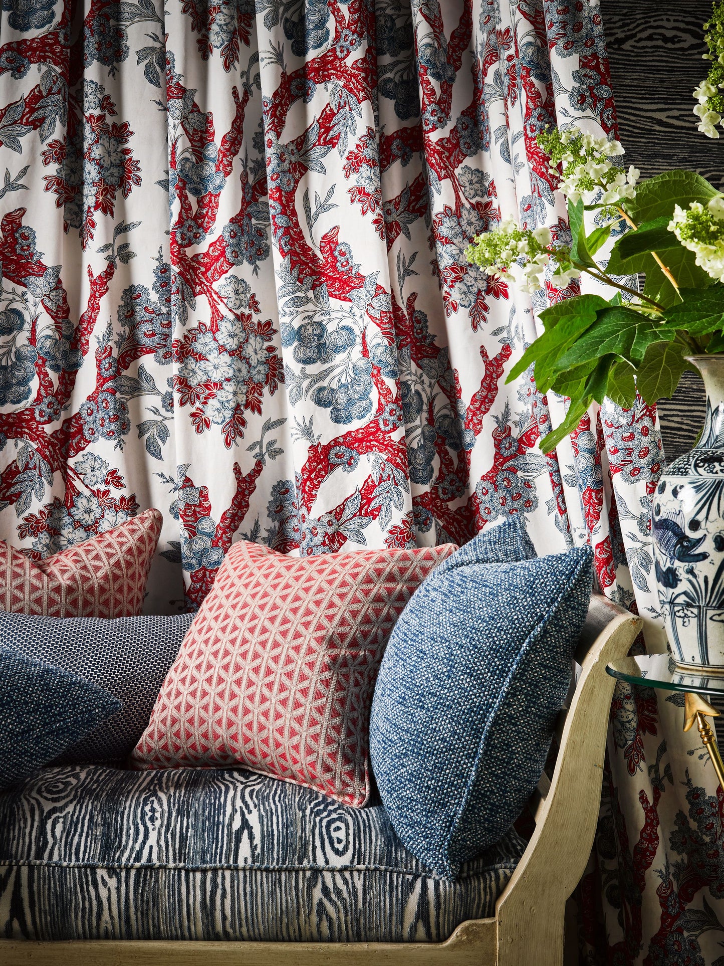 Floral red-blue curtains over a blue patterned sofa with pink diamond cushions and a blue-and-white vase with greenery.