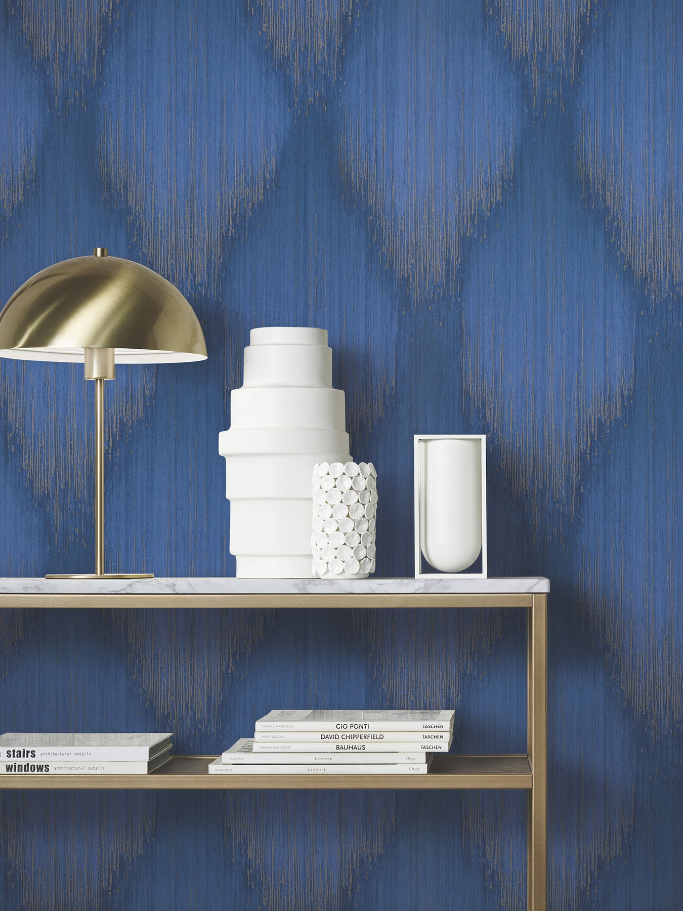 Blue wallpaper with gold pattern, brass dome lamp, white ceramic vases and textured sculpture on marble shelf, stacked books below.