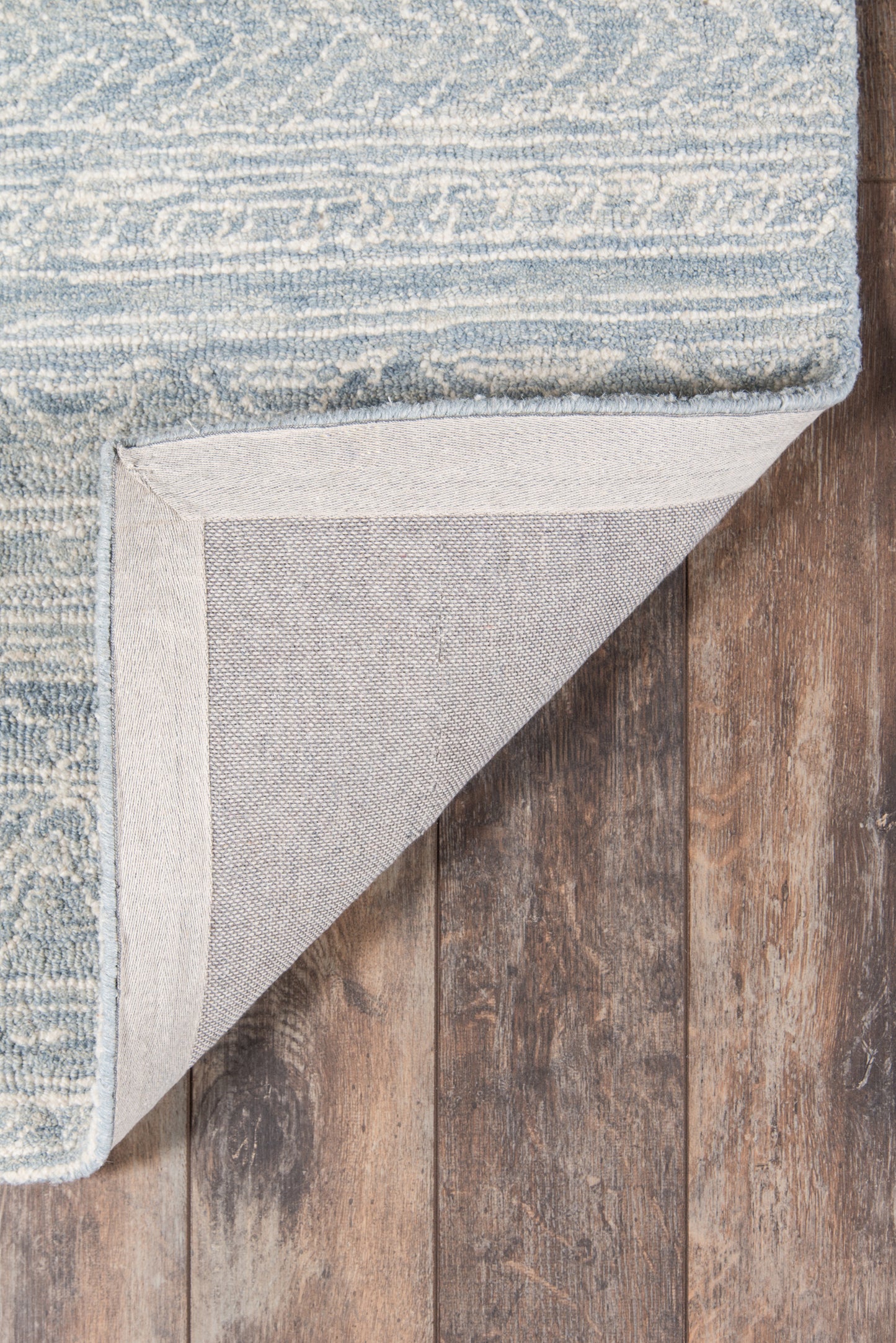 Blue-gray woven rug with subtle striped pattern, light beige edge binding; corner folded to show gray underside on wood floor.