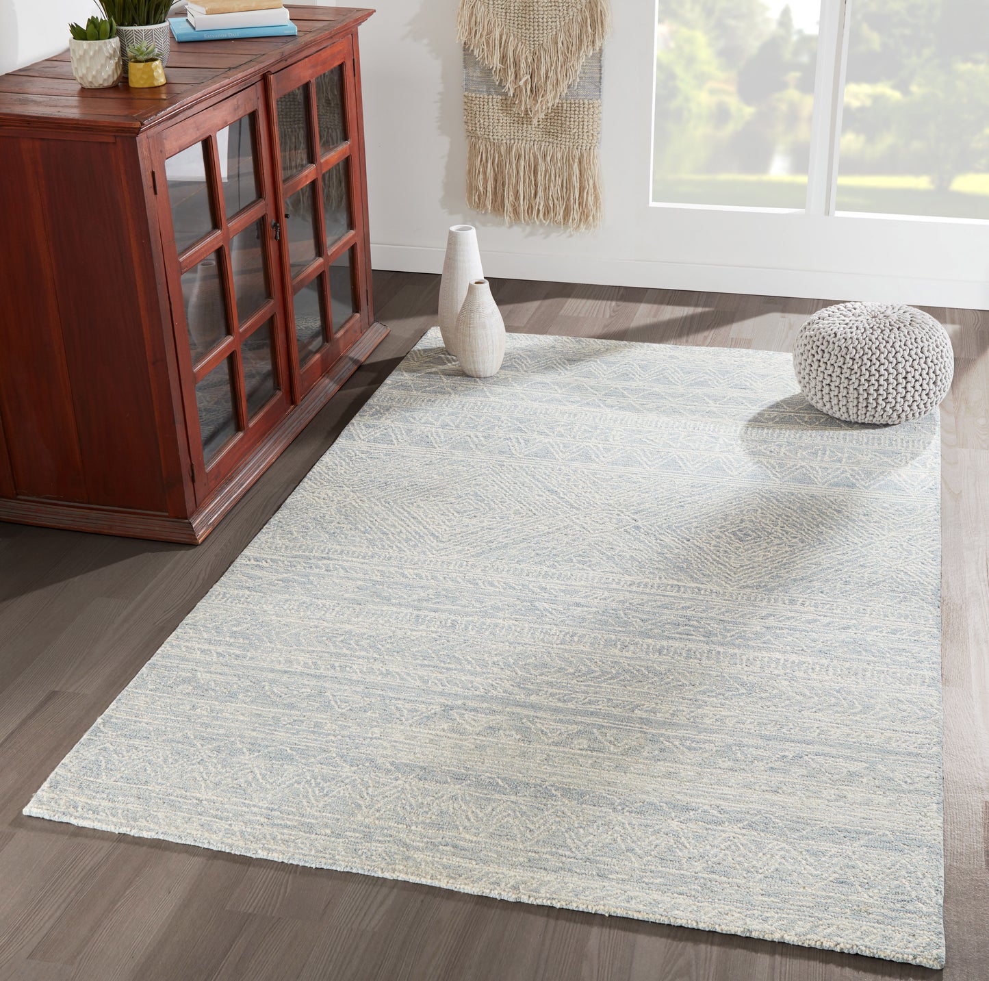 Light blue-gray area rug with subtle geometric pattern in a cozy living room beside a wooden cabinet and knitted pouf.