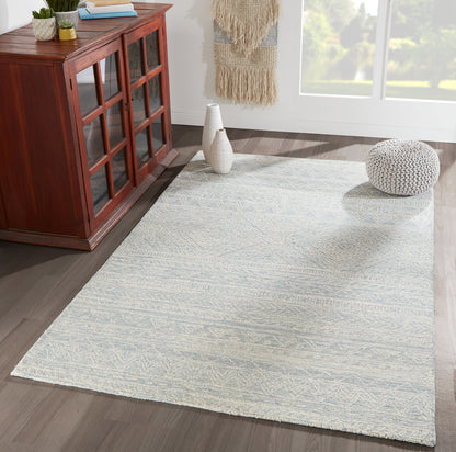Rectangular light gray patterned area rug on dark wood floor beside a reddish-brown cabinet, white vases and a knitted pouf.