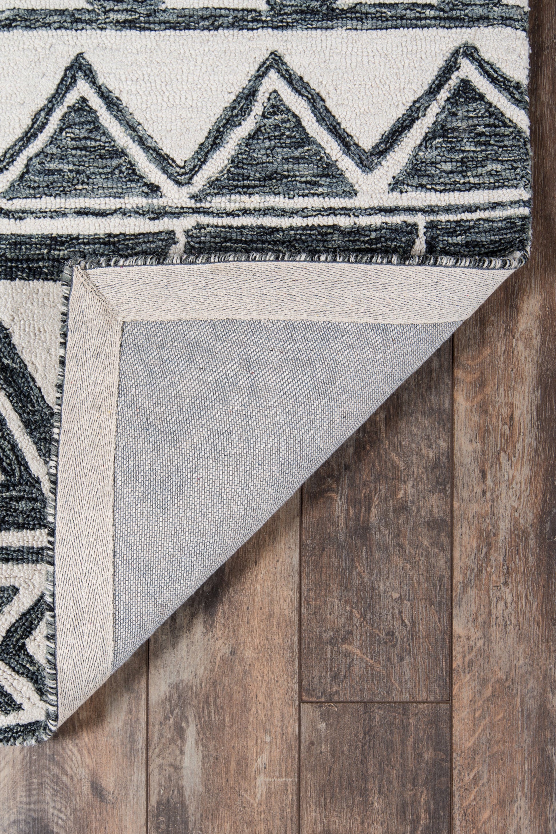 Geometric rug with navy triangle chevron border on light background; folded corner reveals beige backing on wood floor.
