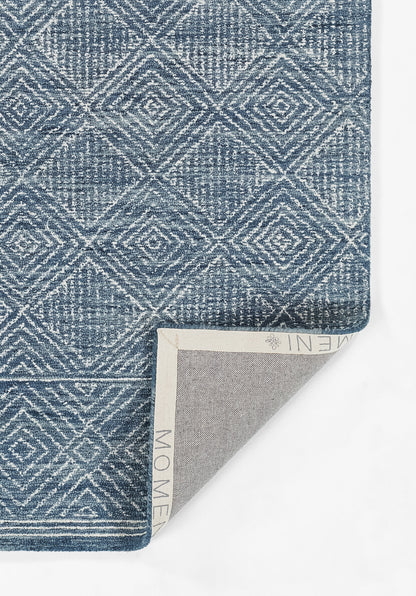 Muted blue geometric diamond rug with white lines, textured weave; folded corner reveals beige backing.