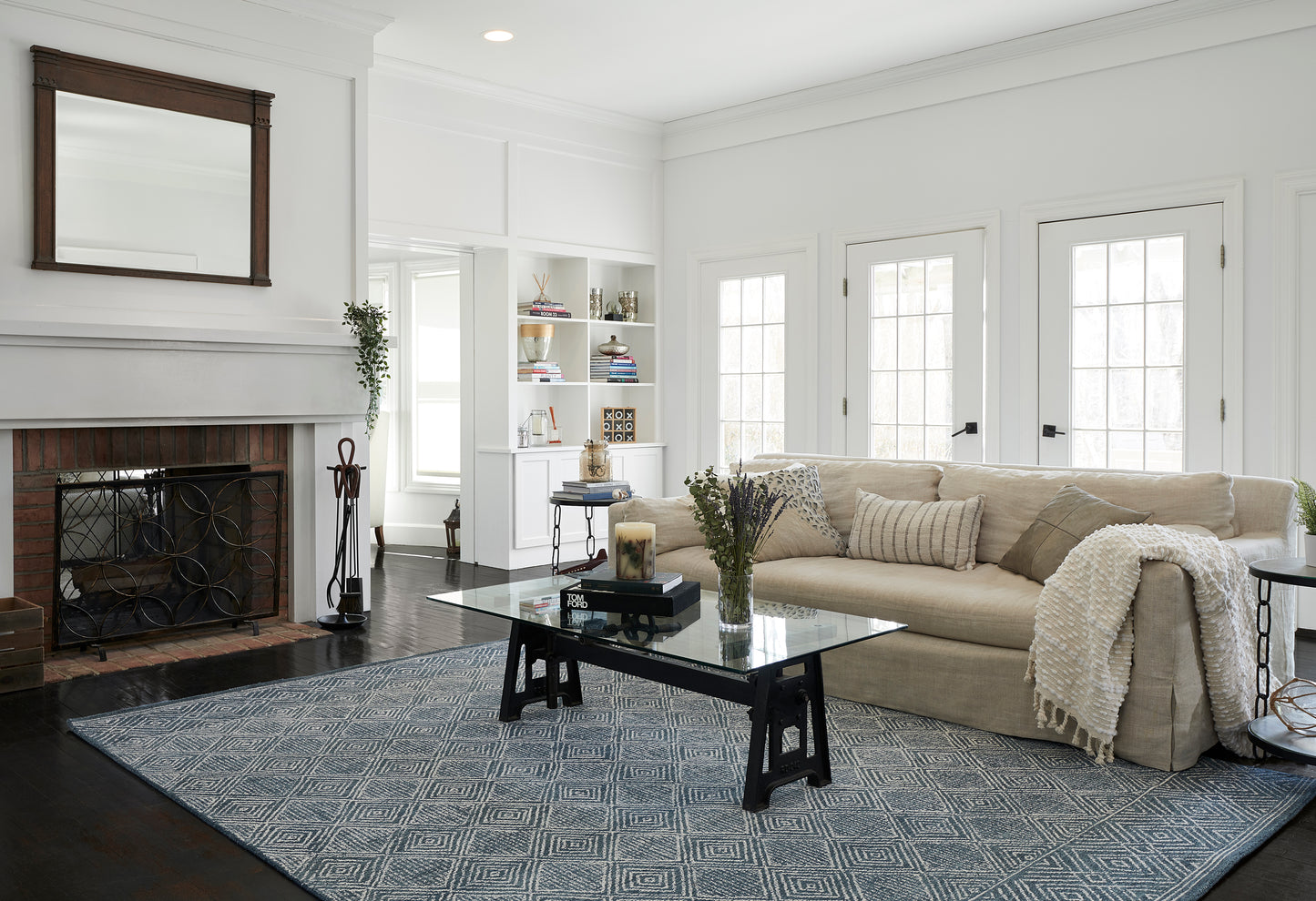 Beige fabric sectional, glass coffee table with black trestle base, blue geometric rug, white fireplace and built-in shelves.
