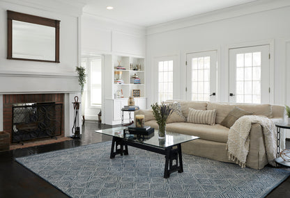 Bright living room: beige sectional, glass coffee table, dark metal legs, blue geometric rug, brick fireplace & white shelves