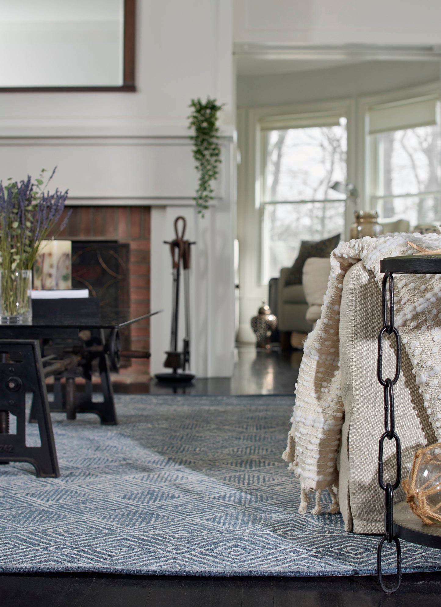 Cozy living room with blue geometric rug, cream knit throw over beige chair, black chain accent, lavender vase on table.