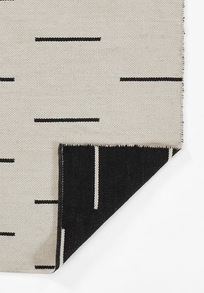 Beige woven rug with black dash line pattern; folded corner reveals black underside with white vertical stripe.
