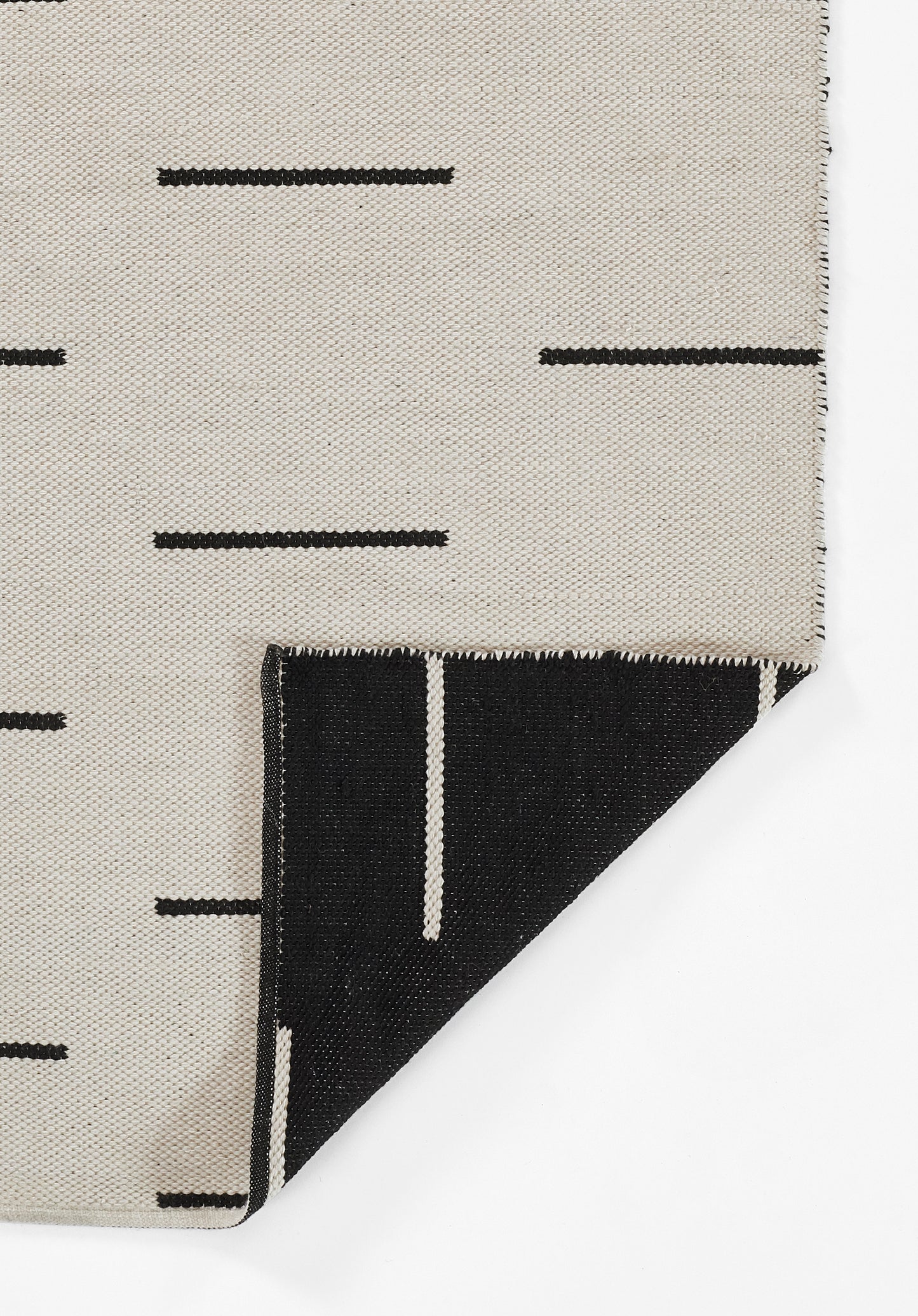 Beige woven rug with minimalist black dash motif; folded corner reveals black reverse with white linear pattern.