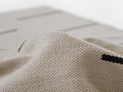 Beige woven rug close-up with a thick texture and a black stripe, draped on a light wooden surface.