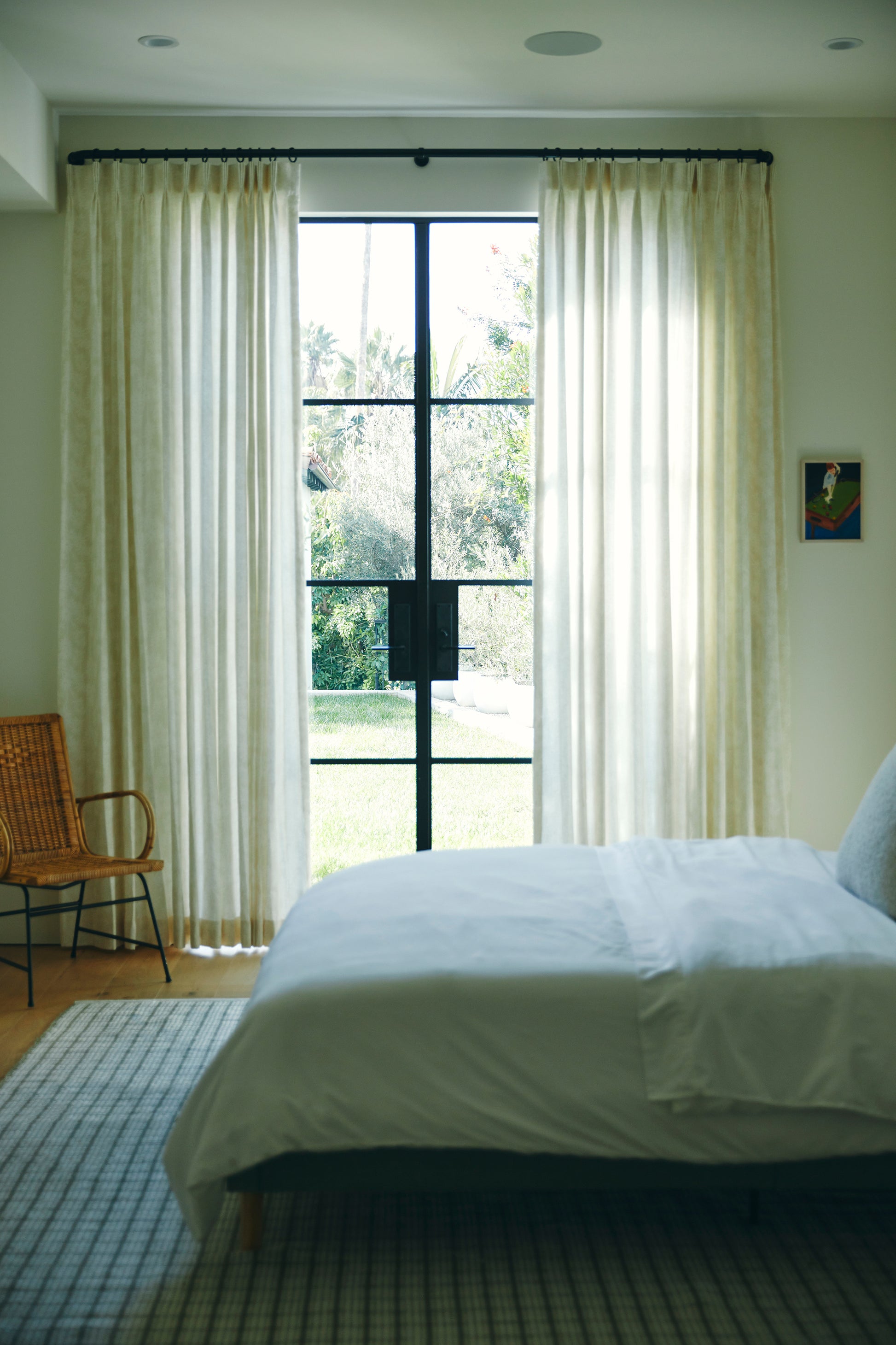 Bright bedroom with white bedding, cream sheer curtains, large black-framed window, wicker chair, and light wood floor.